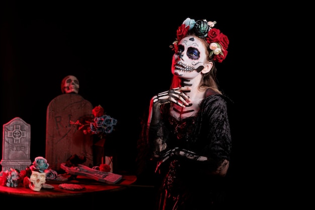 Female model acting glamorous as goddess of dead in studio, wearing flowers crown and black halloween costume. Portraying holy santa muerte or la cavalera catrina, ritual make up.