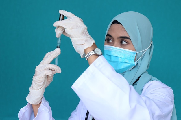 Female Muslim doctor in hijab wearing protective gloves and medical mask holding injection syringe w