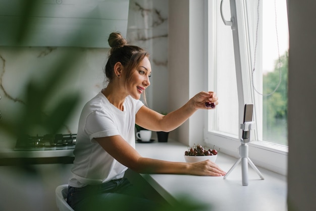 Female nutritionist blogger leads a live broadcast or video conference about the benefits of fresh berries sitting in her kitchen at home