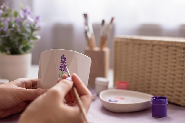 Female paints with a brush a sprig of a lavender flower on a white ceramic pot