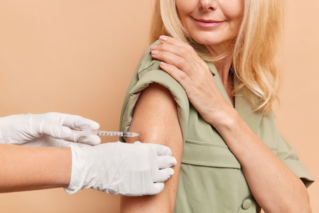 Female patient receives coronavirus vaccine in arm has doctors appointment inn hospital wears dress poses indoor against beige wall