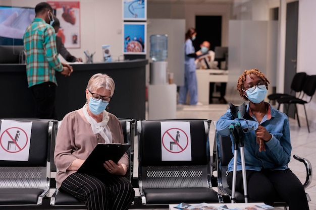 Female patient with impairment sitting in hospital reception lobby, having appointment with medical specialist. Woman with crutches and face mask doing checkup visit, waiting room.