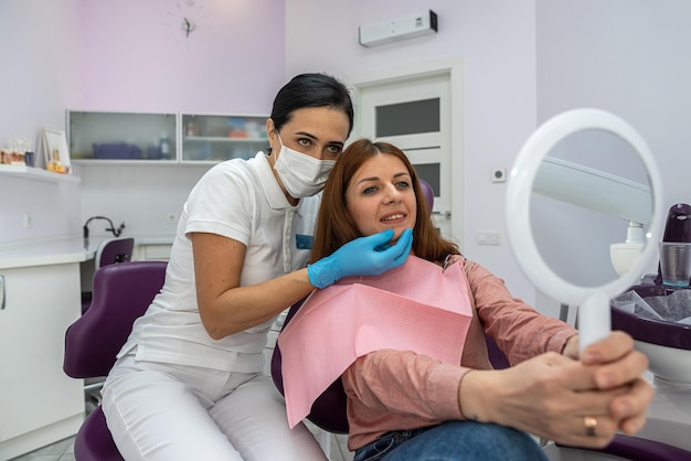 Female patient with a mirror in her hands examines beautiful treated teeth next to doctor