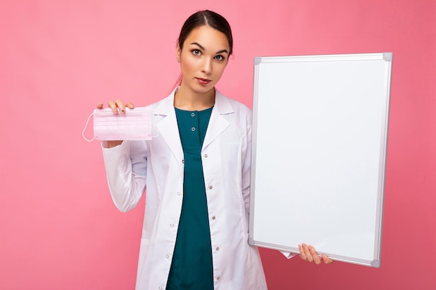 Female person doctor in a white medical coat holding blank board with copy space for text and protective mask isolated on pink background. Epidemic concept