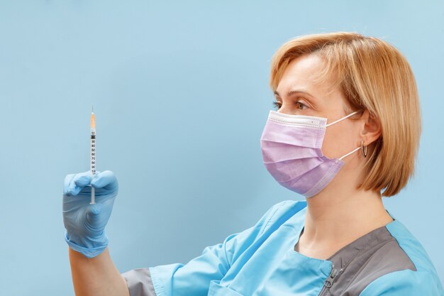 Female physician in a medical uniform, a protective mask and latex gloves holding a syringe with liquid medicine for injection on the blue surface