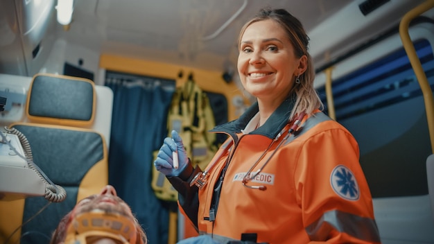 Female Professional EMS Paramedic Ride to Healthcare Hospital with Injured Patient on the Ambulance She Uses Flashlight to Check the Wounds on the Sick Young Man She is Happy and Smiles on Camera