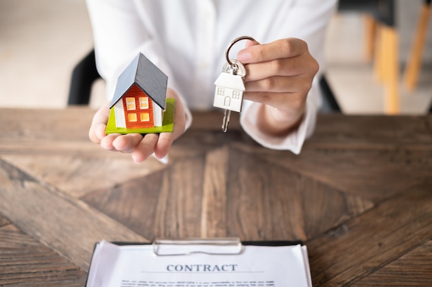 Female real estate agent hand holding house model and house key with contract on table