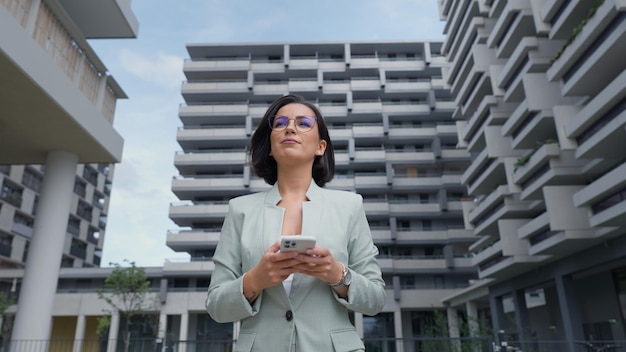 Female realtor wearing formal suit holding smartphone while going through the financial district