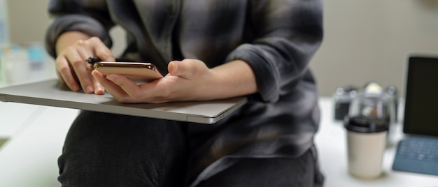 Female sitting on table and using smartphone on laptop on her lap