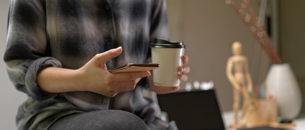 Female sitting on worktable and using smartphone while holding coffee