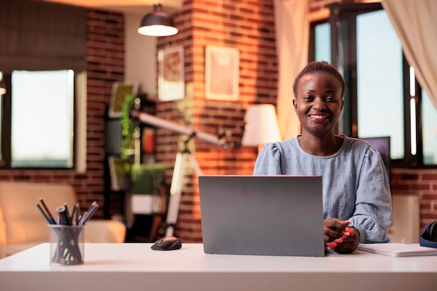 Female smiling freelancer working on laptop in modern home office. Young successful african american businesswoman at workplace in contemporary room with warm sunset light