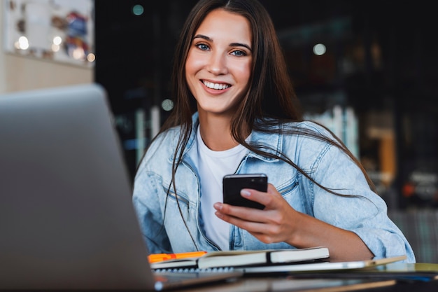 Female smiling, looking at the camera. Attractive business woman sits at table in front of laptop talks on mobile phone