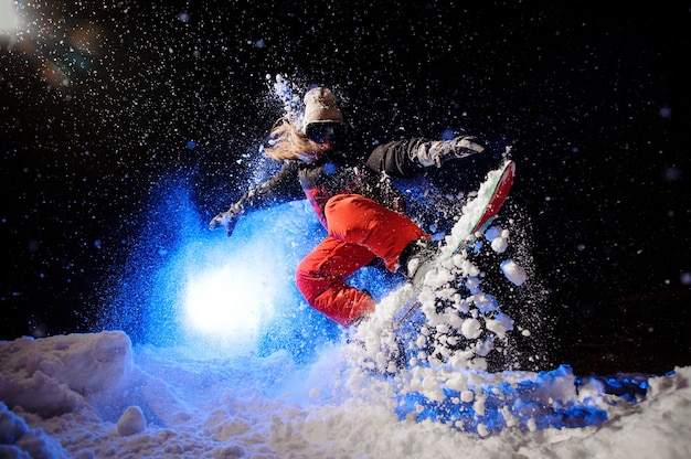 Female snowboarder dressed in a orange sportswear jumping on the mountain slope in the night under the blue light