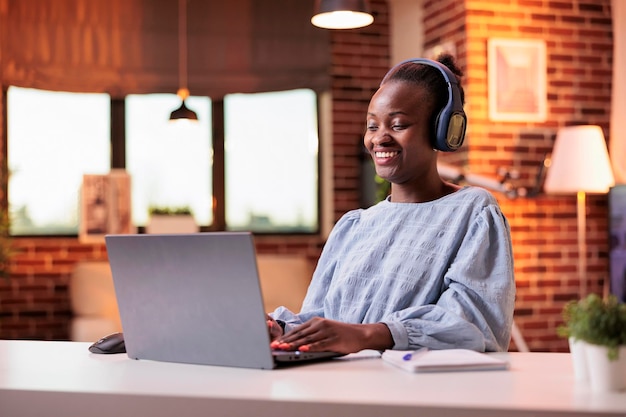 Female student in headphones listening to tutorial on laptop at home. Smiling african american woman watching entertaining video in modern room with beautiful warm sunset light