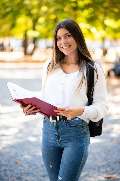 Female student holding a book outdoor in the park and smiling