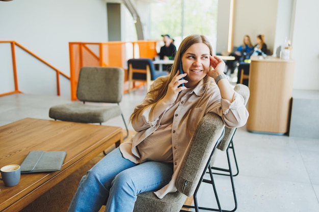 A female student is sitting on a sofa in a bright cafe with coffee and talking on the phone Remote work online in a cozy work cafe