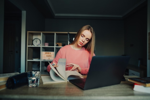 Female student in a pink sweater studies remotely at home looks in a notebook with a serious face