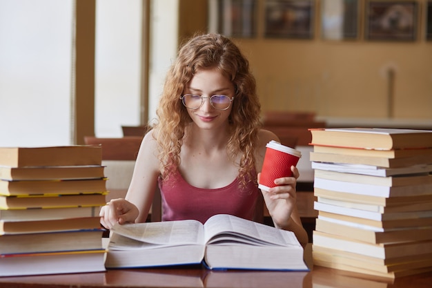 Female student posing with coffee in hand, suraunded with stacks of books at reding room. Young woman sitting at table doing assignments in college library, preparing for classes. Education concept.