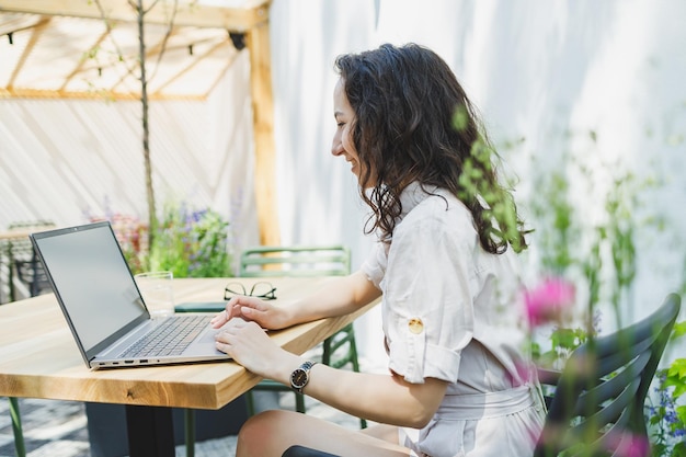 A female student sits on the summer terrace of a cafe and works at a laptop Female freelancer works remotely online while sitting in a summer cafe Remote work