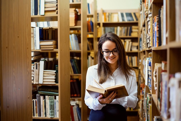 Female student with books in her hands between shelves full of books Smiling girl likes studying and eager to enlarge her knowledge and skills