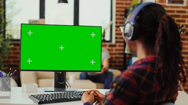 Female student with headset looking at greenscreen display, listening to music on headphones and working with isolated mockup template. Using chroma key copyspace on computer monitor.