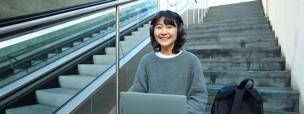 Female student works on project remotely sits on stairs street works on laptop listens music in