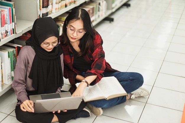 female students finishing assignment together using laptop and book at the library