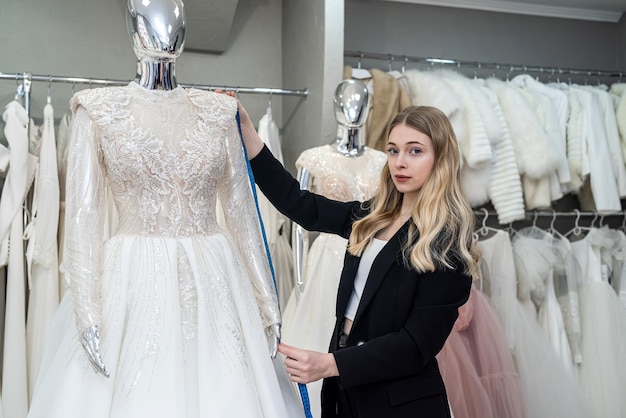 Female tailor measuring elegant wedding dress on mannequin using tape measure