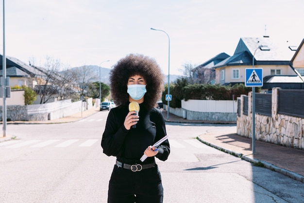 Female television reporter journalist with afro hair, wearing a surgical mask to protect herself from a virus, reporting on a story on the street