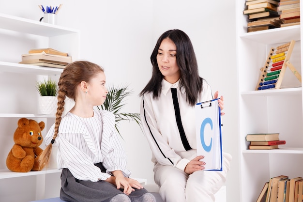 Female therapist working with kid with problem in pronunciation in the white cabinet.