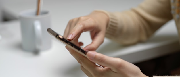 Female touching on smartphone while sitting at white office desk with coffee mug