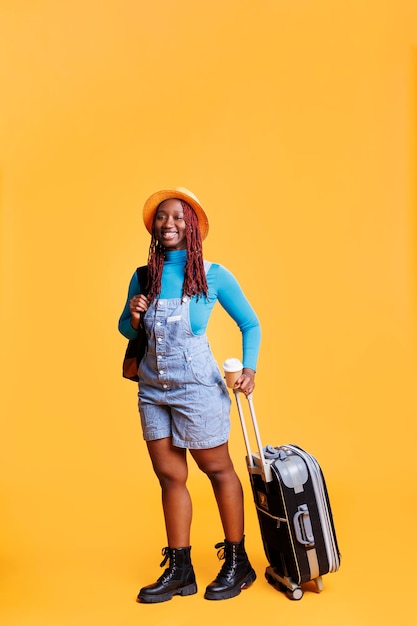 Female traveller drinking cup of coffee and carrying trolley bags, enjoying caffeine refreshment in studio. Young woman serving latte drink for international flight abroad, holiday trip.