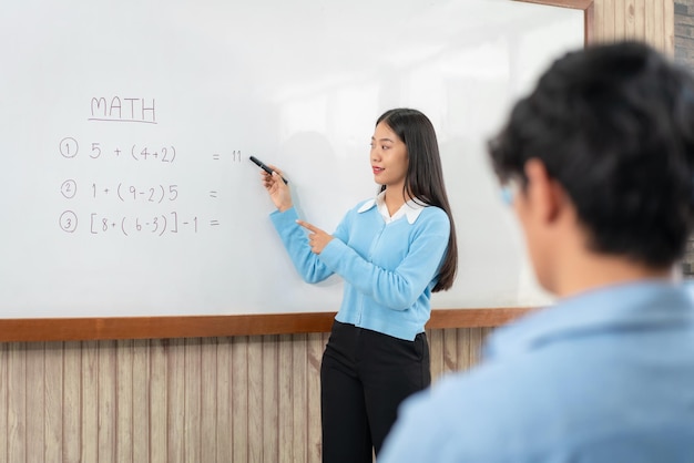 Female tutor standing in front of whiteboard and writing math equations on board to explaining for student during math class in the classroom