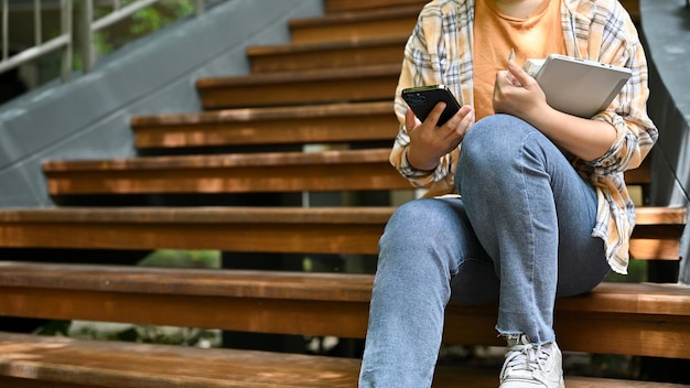 A female uses her smartphone while sitting on the stairs in her campus's park cropped image