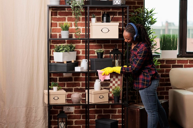 Photo female wearing rubber gloves dusting furniture shelves in modern apartment, while listening music using headphones. tired woman cleaning new home in the city they just moved into.