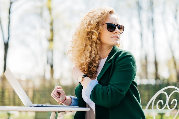 Female with fluffy blonde hair wearing sunglasses sitting on in the sun in front of laptop