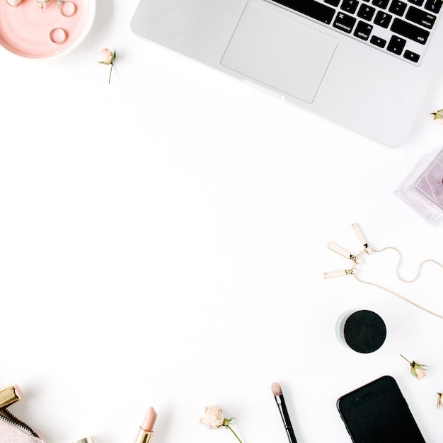 feminine desk workspace with laptop, clutch, cosmetics, phone, sunglasses, lipstick rose buds on white.