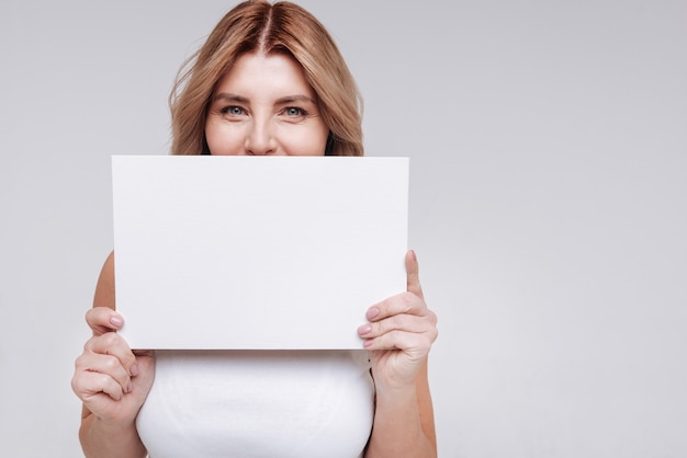 Feminine and strong. Positive hilarious adorable lady using a piece of paper covering up half of her face while posing