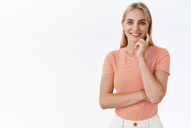 Feminine tender goodlooking woman with blond hair striped tshirt cross one hand and touch cheek lovely gazing camera look trustworthy and confident standing white background