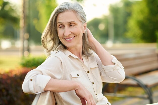 Femininity. Cheerful cute adult blonde woman looking back sitting on park bench on summer day