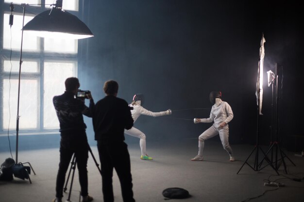 Photo a fencing training in the studio two women in protective costumes having a duel two cameramans standing near the tripod and shooting the duel