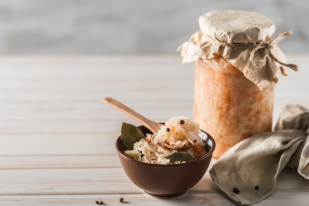 Fermented cabbage in a brown plate and in a glass jar in the background with a wooden spoon and a linen napkin on a light background.