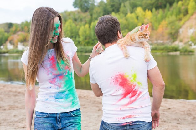 Festival holi, holidays, tourism and nature concept - Couple dressed in white shirts holding cat and covered with colorful dust.