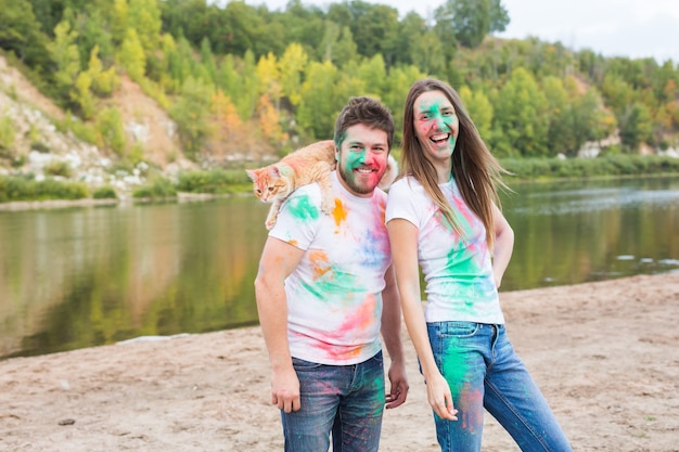 Festival holi, holidays, tourism, pet and nature concept - Portrait of woman and man with cat covered multi-colored dust.