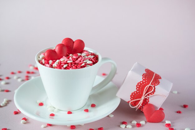 Festive background.  Coffee cup, full of multicolor sweet sprinkles sugar candy hearts and packing Valentine's  Day gifts.  Love and Valentine's day concept.