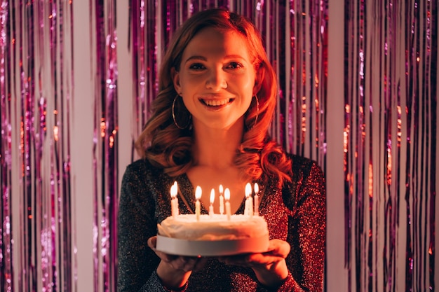 Festive bakery Happy woman Birthday party Smiling inspired lady feeling amazed holding cake with candle lights on shimmering purple cascade curtain background