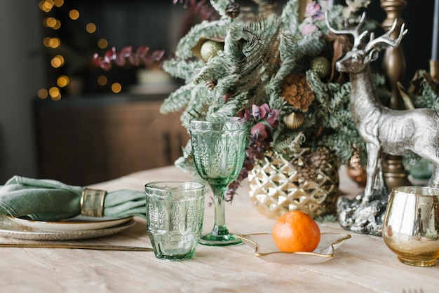 Festive Christmas dinner setting Green wine glasses branches of fir or spruce in a vase plates cutlery and tangerine on a background of bokeh lights