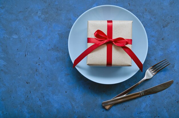 Photo festive concept. gift in craft eco paper with a red ribbon on blue plate with fork and knife on blue background with copy space.