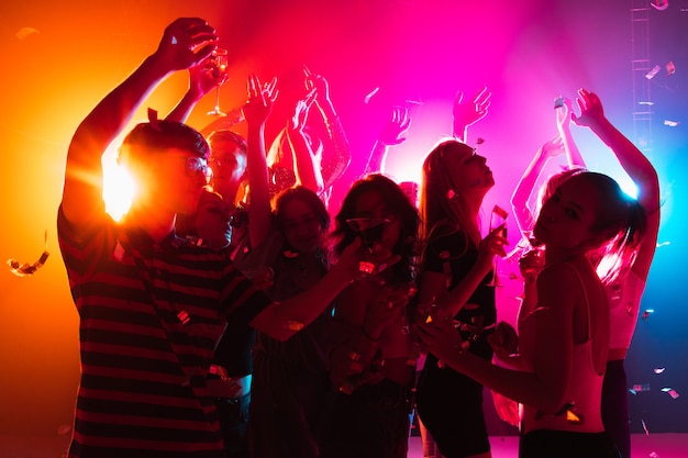 Festive. A crowd of people in silhouette raises their hands, dancing on dancefloor on neon light background. Night life, club, music, dance, motion, youth. Bright colors and moving girls and boys.