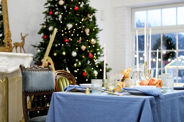 The festive dinner table stands against the backdrop of a Christmas tree On the table there is a blue tablecloth plates glasses and decor